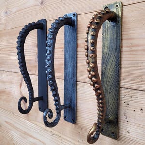 Peut inclure: Trois poignées de porte en métal en forme de tentacules de poulpe. Les poignées sont noires, grises et bronze, chacune fixée sur une plaque arrière rectangulaire. Les tentacules courbent avec des textures détaillées de ventouses.