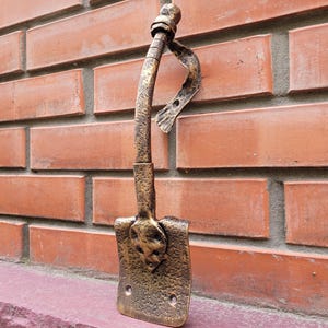 Puede incluir: Un gancho de pared decorativo de metal color bronce con forma de pala pequeña. El gancho está montado en una pared de ladrillo rojo. El extremo en forma de pala tiene un diseño de calavera.