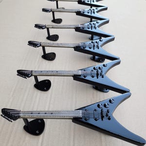 Può includere: Sono esposte sei chitarre elettriche nere con corpo a forma di V. Ogni chitarra è montata su un supporto nero. Le chitarre hanno corde e meccaniche argentate. La finitura lucida riflette la luce, evidenziando i dettagli degli strumenti.