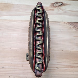 May include: A decorative hot dog sculpture with a bun, sausage, mustard, and ketchup. The bun is brown with gold accents, the sausage is brown, and the condiments are red and gold. The sculpture has a metal bracket for hanging.