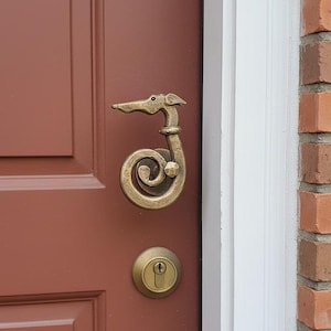 Puede incluir: Un aldaba de puerta de color bronce con forma de cabeza y cuello de perro, con un cuerpo rizado. La aldaba está montada en una puerta de color marrón rojizo, con una cerradura de puerta de color bronce a juego. La puerta está al lado de una pared de ladrillo.