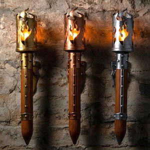 Pode incluir: Três tochas de parede com um design de chama. As tochas têm um cabo de madeira e detalhes metálicos em ouro, cobre e prata. As chamas são iluminadas, criando um brilho quente. As tochas são montadas numa parede de pedra.