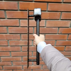 Puede incluir: Un martillo y una llana de alba&ntilde;il de metal negro contra una pared de ladrillos rojos. El martillo tiene una cabeza cuadrada y un mango texturizado. La llana es rectangular con borde dentado.