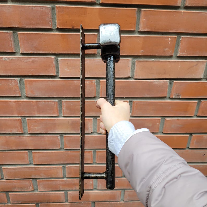 Puede incluir: Un martillo y una llana de alba&ntilde;il de metal negro contra una pared de ladrillos rojos. El martillo tiene una cabeza cuadrada y un mango texturizado. La llana es rectangular con borde dentado.
