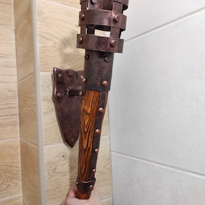 May include: A wall-mounted torch with a wooden handle and a metal cage. The cage is decorated with rivets and has a copper finish. The torch is mounted on a metal bracket.