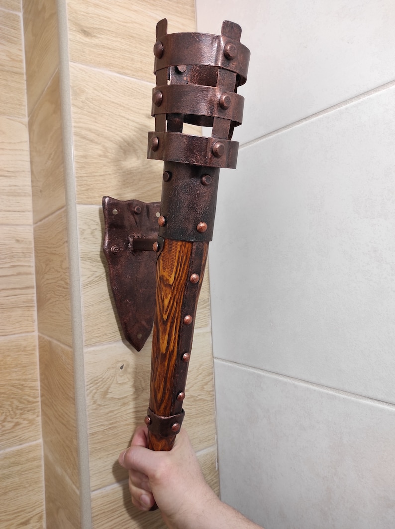 May include: A wall-mounted torch with a wooden handle and a metal cage. The cage is decorated with rivets and has a copper finish. The torch is mounted on a metal bracket.