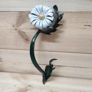 May include: A white daisy with a gold centre is attached to a green metal stem. The stem is curved and has leaves. The metal is painted green and gold.