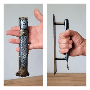 May include: A decorative door handle with a fish design. The handle is a combination of silver, bronze, and gold colors. The fish-shaped handle is attached to a rectangular plate. The handle is held in a hand.