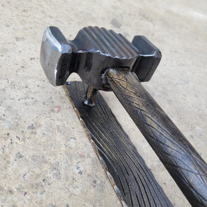Puede incluir: Un martillo artesanal de metal oscuro con un mango texturizado y una base rectangular. La cabeza del martillo tiene una superficie de golpe plana y una pena curva. El mango tiene un dise&ntilde;o de vetas de madera. El martillo probablemente se utiliza para trabajos en metal o construcci&oacute;n.