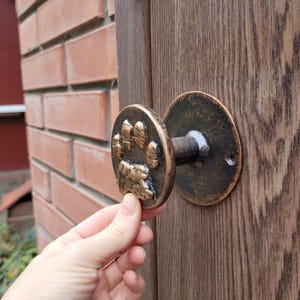 May include: Bronze door knob featuring a paw print design. The round knob has a detailed paw print in the centre. It is attached to a wooden door. The background includes a brick wall.
