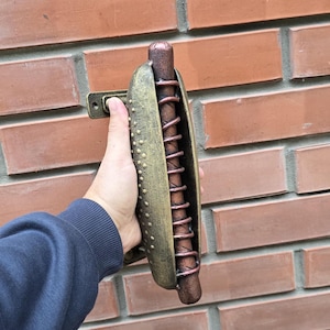 Pode incluir: Uma maçaneta de porta de ouro e cobre em forma de cachorro-quente com um design em espiral. A maçaneta está montada em uma parede de tijolo.