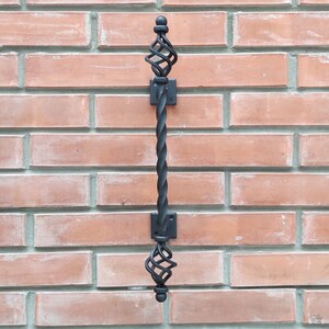 May include: Black metal door handle with a twisted design and decorative finials. The handle is mounted on a brick wall.