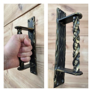 May include: Black and gold decorative door pull. The handle features a twisted design and is mounted on a rectangular plate with decorative edges. The door pull is shown mounted on a wooden surface.