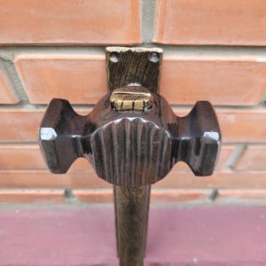 Puede incluir: Un martillo artesanal marr&oacute;n oscuro con cabeza y mango texturizados. El martillo est&aacute; montado sobre un soporte de madera con un acento dorado. El fondo es una pared de ladrillos.