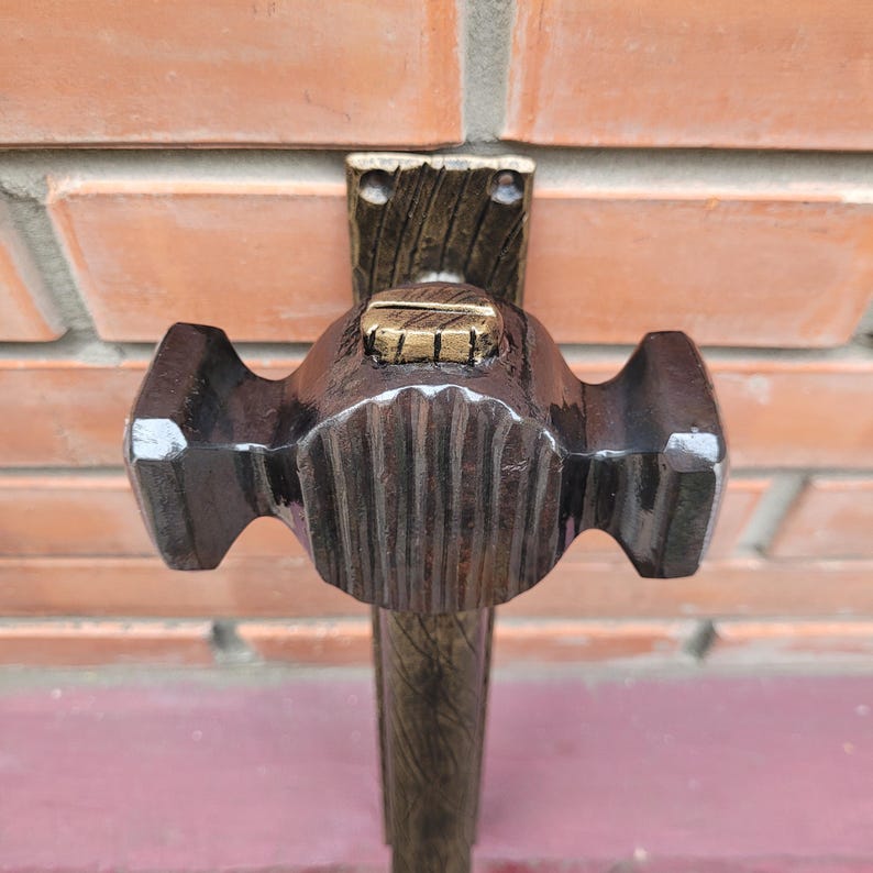 Puede incluir: Un martillo artesanal marr&oacute;n oscuro con cabeza y mango texturizados. El martillo est&aacute; montado sobre un soporte de madera con un acento dorado. El fondo es una pared de ladrillos.