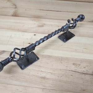 May include: A black metal handrail with a twisted design and rounded ends. The handrail is mounted to a wall with two rectangular brackets.