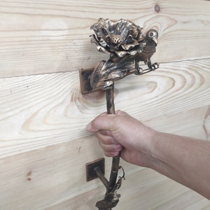 May include: A decorative door handle made of metal, shaped like a flower with a bird perched on it. The handle is painted in a gold finish and has a rustic look.