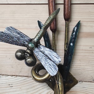 May include: A metal dragonfly sculpture with a blue and silver finish, perched on a set of three metal reeds. The reeds are painted in shades of green and brown.