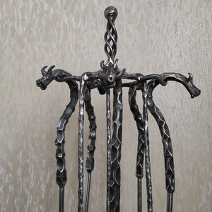 Pode incluir: Um conjunto de ferramentas para lareira em ferro forjado com tema de dragão. O conjunto inclui um suporte com detalhes de cabeça de dragão e várias ferramentas. O suporte tem um topo torcido e as ferramentas têm cabos texturizados. O metal é cinza escuro.