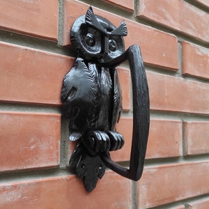 May include: Black metal owl door knocker with a detailed design. The owl is perched on a branch with its wings spread. The knocker has a ring handle for knocking.