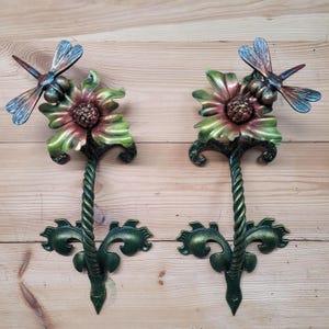 Op de afbeelding: Twee decoratieve metalen wandkandelaars. Elke kandelaar heeft een libelle bovenop een bloem met groene en rode bloemblaadjes. De kandelaars hebben een gedraaide steel en decoratieve groene bladeren. De libellen vleugels zijn blauw en brons.