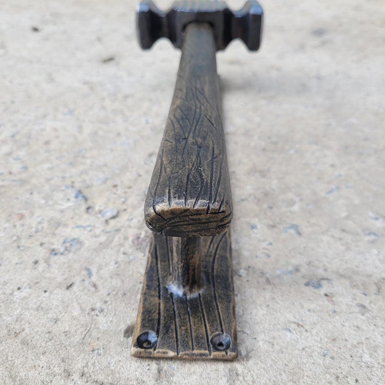 Puede incluir: Un tirador de puerta de estilo r&uacute;stico, color bronce, con dise&ntilde;o de martillo. El mango tiene un aspecto texturizado de veta de madera y la base presenta dos orificios para tornillos. La longitud total es de unos 30 cm.