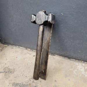 Puede incluir: Un martillo de madera hecho a mano con un acabado oscuro. La cabeza del martillo es rectangular con una superficie texturizada, y el mango est&aacute; hecho de dos piezas de madera paralelas con detalles de vetas talladas. El martillo est&aacute; dise&ntilde;ado para fines decorativos o de exhibici&oacute;n.