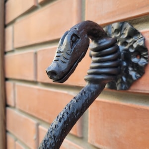 Könnte beinhalten: Ein schwarzer, dekorativer Türklopfer in Form einer Schlange. Der Klopfer ist aus Metall gefertigt und an einer roten Backsteinmauer befestigt. Der Schlangenkopf ist mit Schuppen und einer strukturierten Oberfläche versehen.