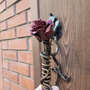 May include: A handcrafted, wrought iron door knocker featuring a burgundy rose with green leaves. The stem is wrapped with a golden vine design. The door knocker is mounted on a wooden door, with a brick wall in the background.
