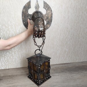 May include: A metal hand holding a lantern. The hand is attached to a wall with a metal axe behind it. The lantern is hanging from the hand by a chain.