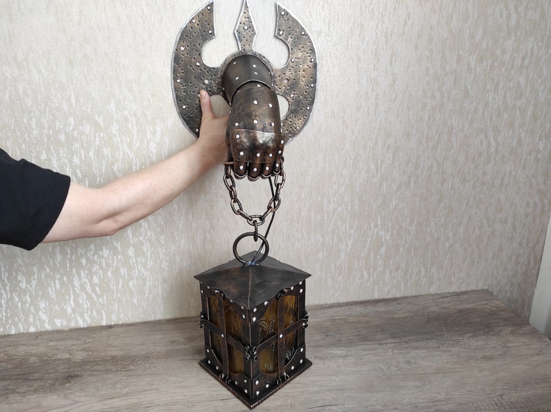 May include: A metal hand holding a lantern. The hand is attached to a wall with a metal axe behind it. The lantern is hanging from the hand by a chain.