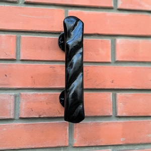 May include: Black metal door handle with a twisted design. The handle is mounted on a brick wall.