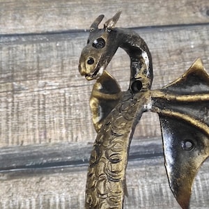 Op de afbeelding: Een gouden en zwarte draken deurgreep met ingewikkelde details. De greep is gevormd als een drakenkop en -nek, met vleugels die aan beide kanten uitsteken. De greep is gemaakt van metaal en heeft een rustieke afwerking.