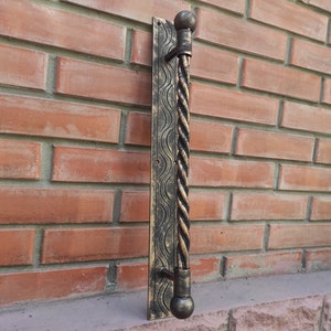 May include: A black metal door handle with a twisted design and gold accents. The handle is mounted on a brick wall.