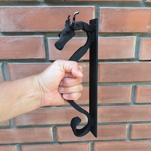 Puede incluir: Pomo de puerta de metal negro con diseño de dragón. El pomo está montado en una pared de ladrillo.
