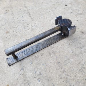 Puede incluir: Un tirador de puerta artesanal de metal oscuro con un dise&ntilde;o en forma de martillo. El mango presenta un patr&oacute;n texturizado de vetas de madera, y la base tiene una serie de l&iacute;neas curvas. La cabeza del martillo es cuadrada y tiene una superficie texturizada. El tirador de puerta est&aacute; dise&ntilde;ado para montarse en una puerta.