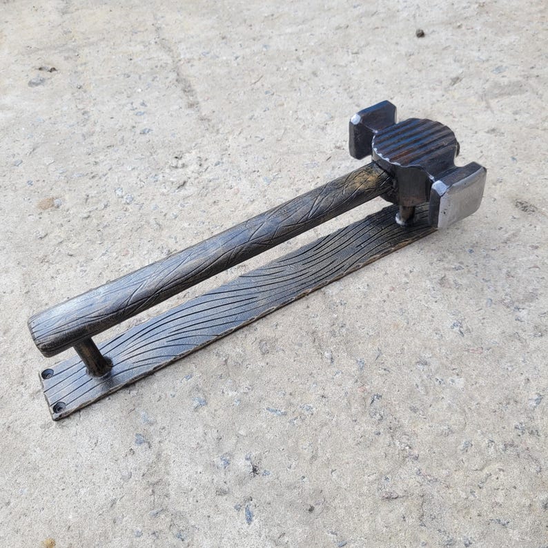 Puede incluir: Un tirador de puerta artesanal de metal oscuro con un dise&ntilde;o en forma de martillo. El mango presenta un patr&oacute;n texturizado de vetas de madera, y la base tiene una serie de l&iacute;neas curvas. La cabeza del martillo es cuadrada y tiene una superficie texturizada. El tirador de puerta est&aacute; dise&ntilde;ado para montarse en una puerta.