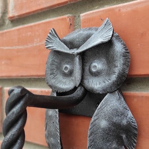May include: A black metal owl door knocker with a twisted metal handle. The owl is mounted on a brick wall.