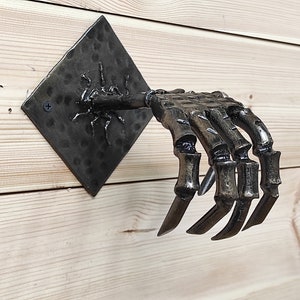 May include: A black metal skeleton hand wall hook with a square metal base. The hand is outstretched with fingers curled.