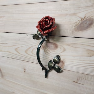 May include: A red metal rose with green leaves and a stem. The rose is attached to a wall.