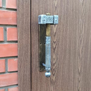 Puede incluir: Una manija de puerta plateada con forma de hacha medieval con un mango de madera. La manija está montada en una puerta de madera.