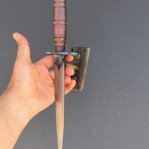 May include: A dagger with a wooden handle and a metal blade. The handle is brown with brass accents. The blade is silver and tapers to a point. The dagger is held against a dark gray background.