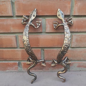 Puede incluir: Dos tiradores de puerta de metal de color dorado con forma de lagartos y superficies texturizadas. Los tiradores están montados en una pared de ladrillo.