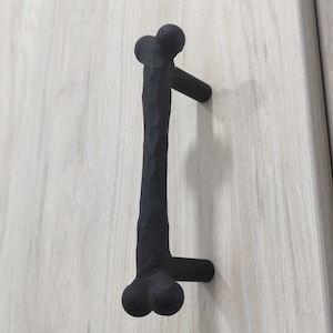 May include: Black metal cabinet pull with a bone-shaped design. The pull has a rough, hammered texture.