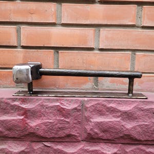 Puede incluir: Un tirador de puerta de metal hecho a mano con un extremo en forma de martillo y un mango texturizado. El tirador de la puerta es de color marr&oacute;n oscuro con una cabeza de martillo plateada y est&aacute; montado sobre una base rectangular.