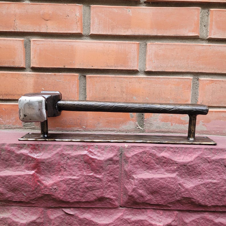 Puede incluir: Un tirador de puerta de metal hecho a mano con un extremo en forma de martillo y un mango texturizado. El tirador de la puerta es de color marr&oacute;n oscuro con una cabeza de martillo plateada y est&aacute; montado sobre una base rectangular.