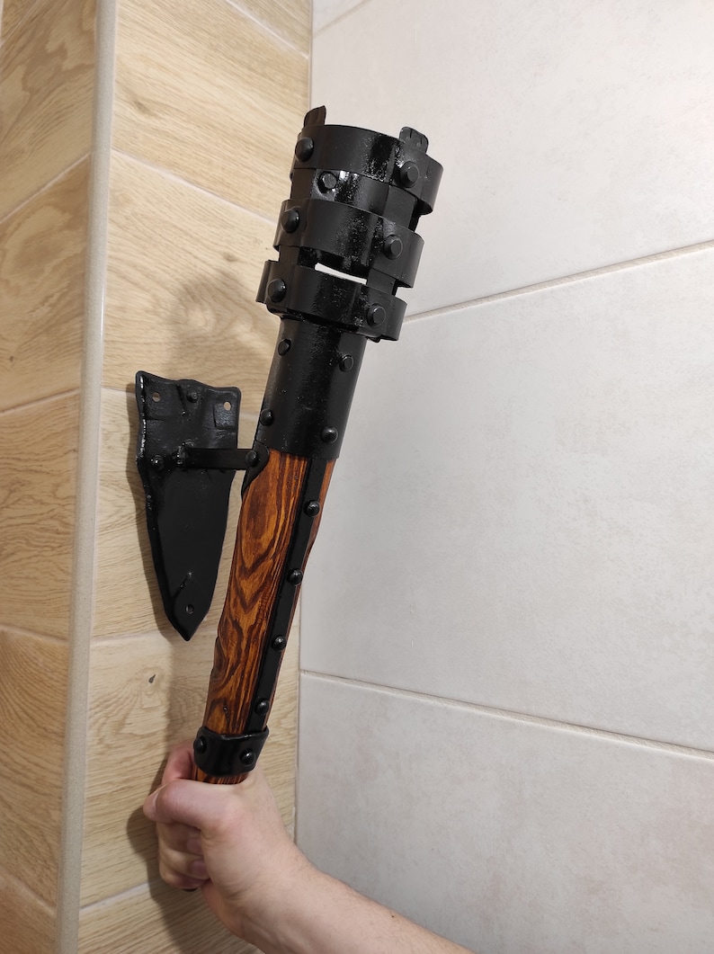 May include: A black metal wall-mounted torch with a wooden handle. The torch has a decorative metal design with rivets and a black finish. The handle is made of wood with a brown stain.