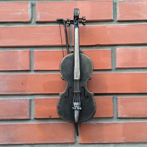 May include: A black and gold metal violin with a bow hanging on a brick wall.