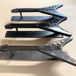 May include: Three metal drawer pulls shaped like V-style electric guitars, each mounted on a rectangular base plate with a wood grain texture. The set includes one silver-toned, one glossy black, and one bronze-toned pull, each featuring strings.