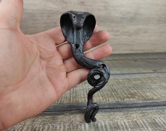 Può includere: Uncino in metallo nero a forma di cobra. L'uncino ha la forma di un cobra con la testa alzata e la coda avvolta attorno al corpo. L'uncino è in metallo e ha una finitura nera.